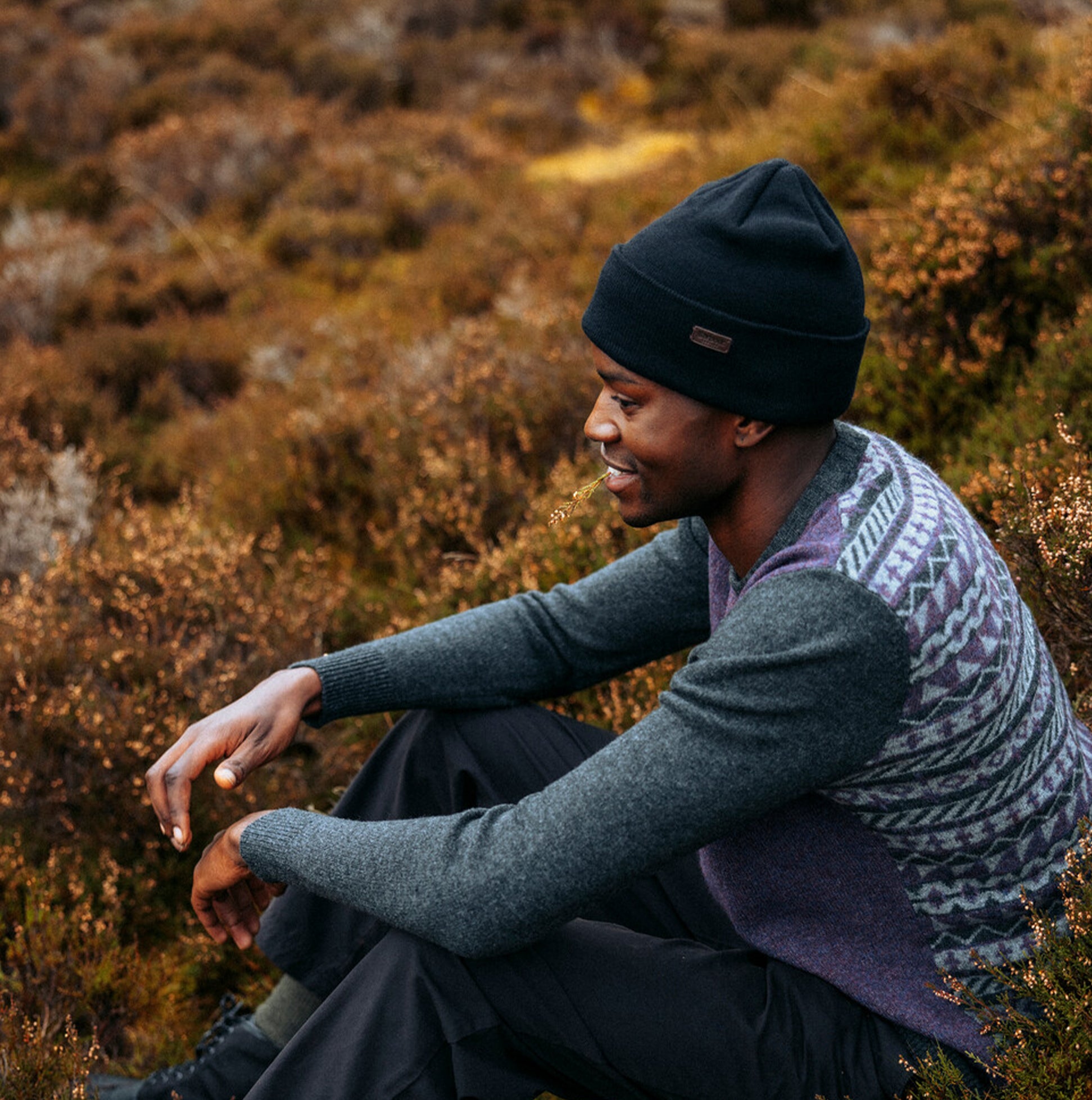 Barbour HEALEY Mens Beanie Black worn by a man sitting in nature, side view, featuring a knitted design.