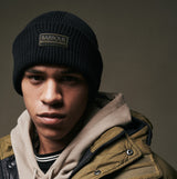Barbour FLEX Mens Beanie Black, front view on model wearing an olive green jacket, showcasing the ribbed design and logo.
