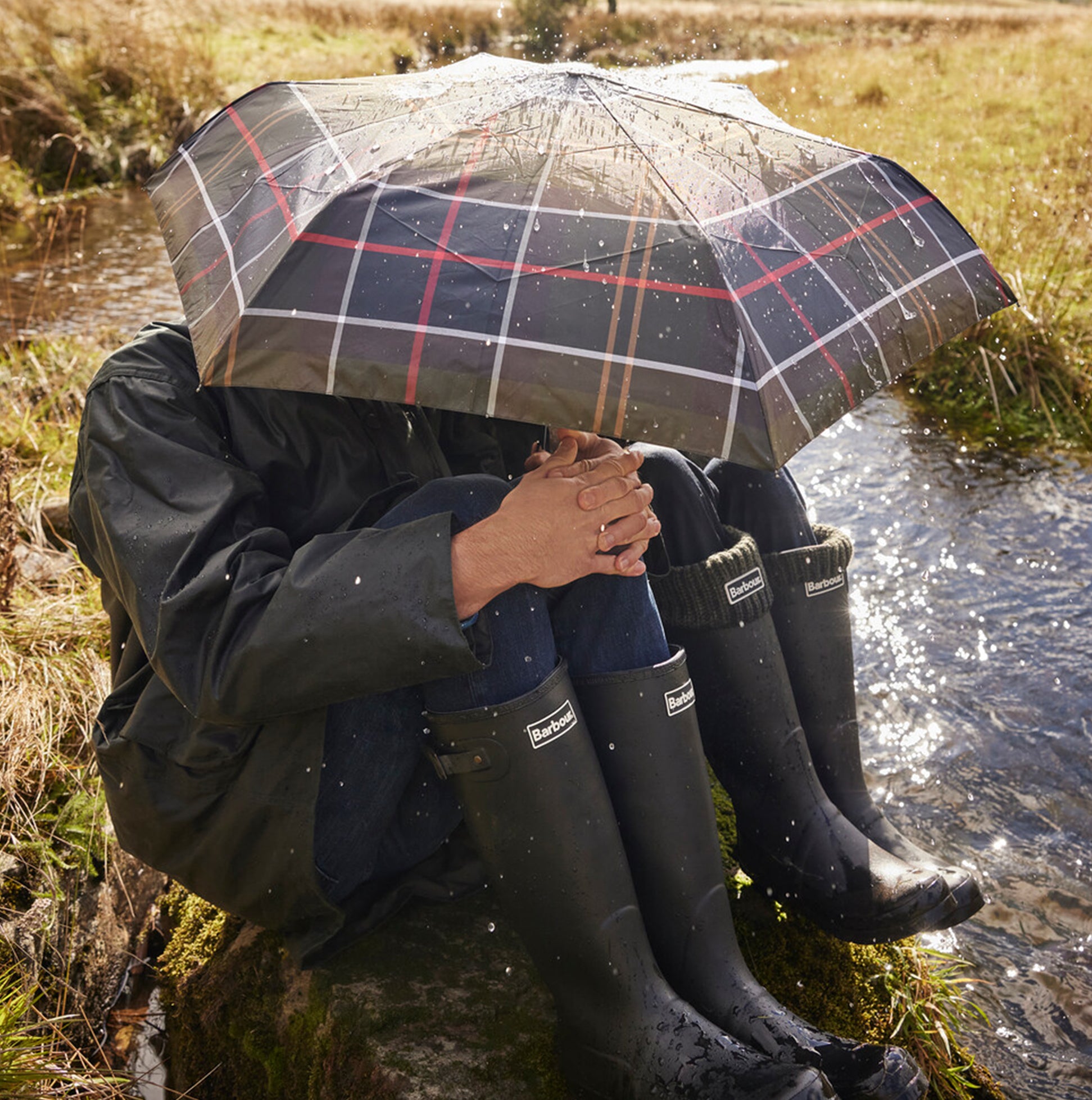 Barbour TARTAN FULL LENGTH Classic Umbrella overhead view, with person sitting beside a stream in waterproof boots.